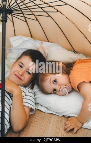 Children in pajamas playing at home lying on the floor under an umbrella, hidden from their parents. Resting on heating floor. Boy and girl posing for Stock Photo