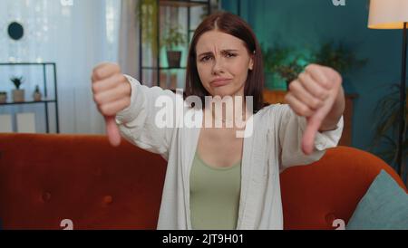 Dislike. Upset girl showing thumbs down sign gesture, expressing discontent, disapproval, dissatisfied bad work at modern home apartment indoors. Displeased young woman in living room sitting on sofa Stock Photo