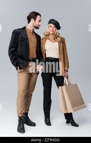 bearded man holding hands with blonde woman in stylish beret standing with shopping bags on grey,stock image Stock Photo