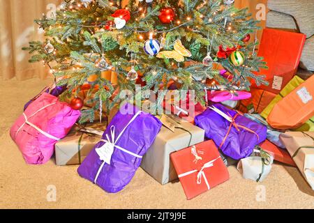 Eco friendly colourful recyclable wrapping paper for Christmas gift & present wrap placed at base of decorated artificial Xmas tree with LED lights UK Stock Photo