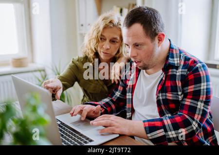 Young man with Down syndrome sitting at desk in office and using laptop, his mother helping him. Stock Photo
