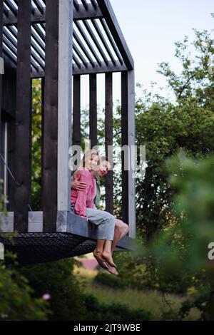 Young couple sitting and cuddling in hammock terrace in their new home in tiny house in woods, sustainable living concept. Stock Photo