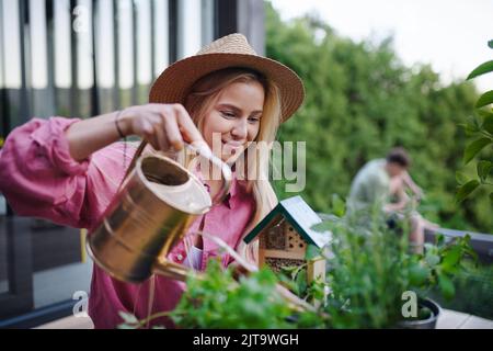 Young woman taking care of her herbs, watering them on terrace in tiny house, sustainable living. Stock Photo