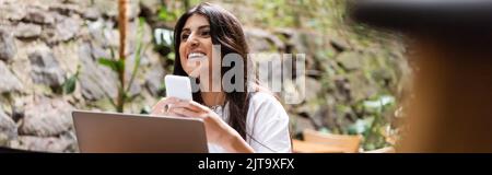 Happy woman holding smartphone near laptop in outdoor cafe, banner,stock image Stock Photo