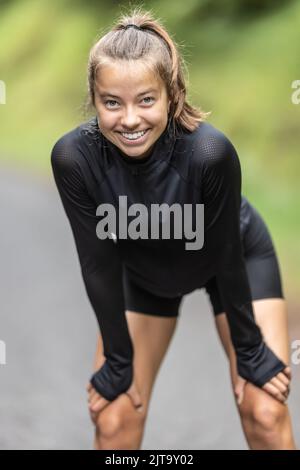 Beautiful girl dressed in black sportswear smiles as she rests after sporting activity outdoors. Stock Photo