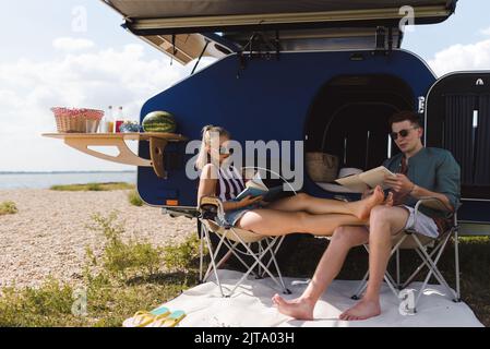 Young couple sitting together in front of van, camping and reading book. Stock Photo
