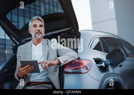 Businessman using tablet while charging car at electric vehicle charging station, closeup. Stock Photo