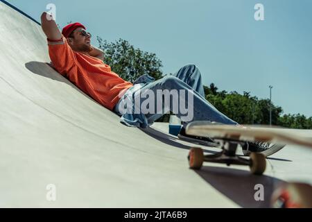 low angle view of happy and stylish man lying on ramp near blurred skateboard Stock Photo