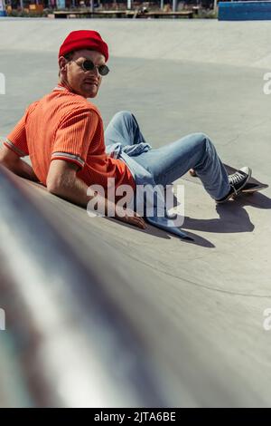 fashionable skateboarder looking at camera while sitting on ramp on blurred foreground Stock Photo