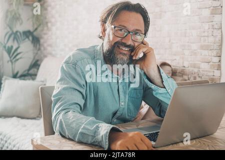 Portrait of cheerful man smiling and looking on camera with computer on the table. Adult young male sitting at the work place at home. People enjoying Stock Photo