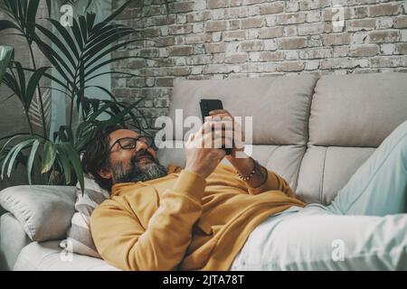 One mature man use mobile phone cellular laying on tne sofa at home in relax leisure online activity. People using cell smartphone indoor in apartment Stock Photo