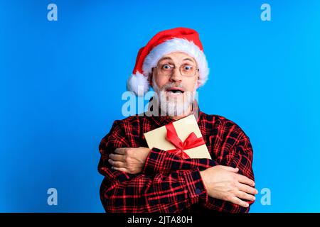 an elderly and well-groomed man in a plaid shirt and santa hat in blue studio background Stock Photo