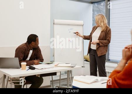 Mature woman making notes on flipchart to present her report for teacher and other students during lesson at classroom Stock Photo