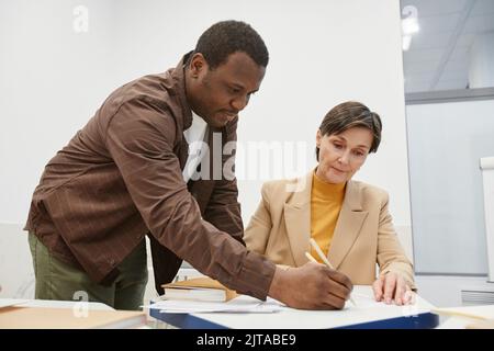 Mature woman talking to her teacher sitting at desk while he explaining her new topic during lesson Stock Photo