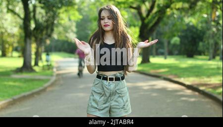 Annoyed confused millennial girl raising arms in disbelief in outdoor park. Puzzled young woman reaction Stock Photo