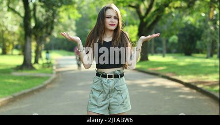 Annoyed confused millennial girl raising arms in disbelief in outdoor park. Puzzled young woman reaction Stock Photo