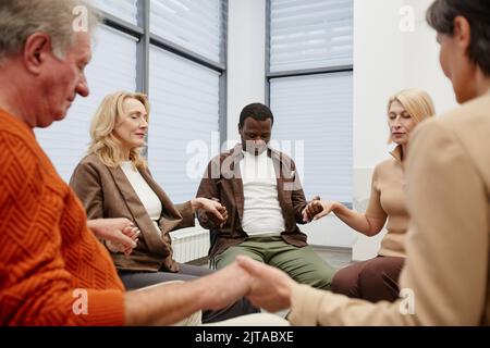 Group of people sitting in circle and holding hands with their eyes closed during psychotherapy Stock Photo