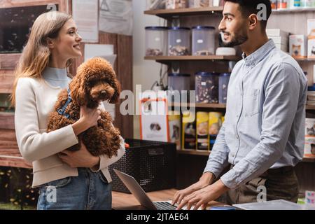 Side view of smiling woman holding poodle near arabian seller in pet shop Stock Photo