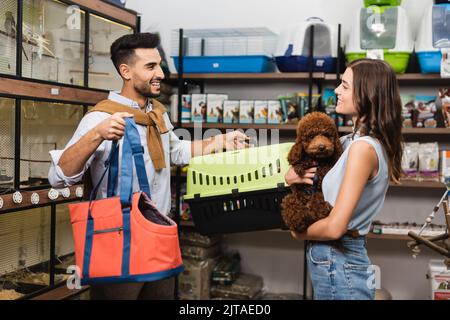 Side view of cheerful interracial couple holding bag and poodle in pet shop Stock Photo
