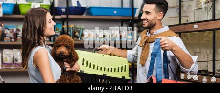 Side view of cheerful interracial couple holding poodle and animal cage in store, banner Stock Photo