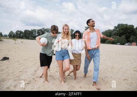 excited multicultural friends walking on sand beach with ball Stock Photo
