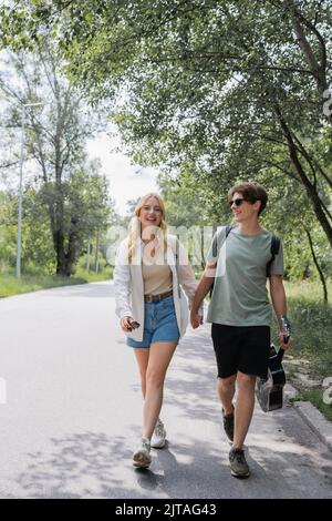 full length of young travelers holding hands and walking on road along forest Stock Photo