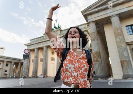 curly tourist with backpack waving hand near brandenburg gate in berlin,stock image Stock Photo