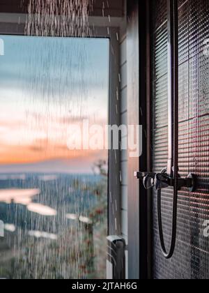 Bathing room interior with panoramic window. Splashes and drops of water from shower in bathroom with panoramic window in summer nature background at sunset. Stock Photo