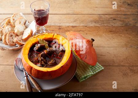 From above delicious meat goulash served in pumpkin near plate with bread pieces and glass of red wine on timber table during lunch Stock Photo