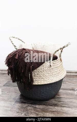 Handwoven seagrass belly or blanket basket with handles with beige and brown throws. Trendy natural multifunctional container for storage laundry, grocery, toy, plant in home. Side view, copy space Stock Photo