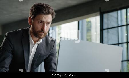 Serious 40s bearded businessman entrepreneur employee standing in office looking at papers report information papers check data in online net service Stock Photo