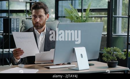Serious adult 40s bearded pensive thoughtful businessman working at computer in office with paperwork. Professional employee worker typing thinking Stock Photo