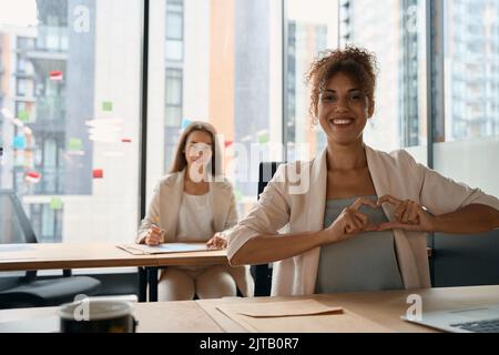 Happy woman shows heart with her hands, sitting in office Stock Photo
