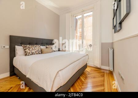 Bedroom with gray fabric upholstered headboard, matching cushions and pillows, large balcony with white windows and shutters, matching blankets, origi Stock Photo