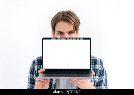 Amazed excited caucasian young man peeking out from behind laptop, looks surprised at camera, stands on isolated white background, holds an open laptop with blank white screen, copy space, mock-up Stock Photo