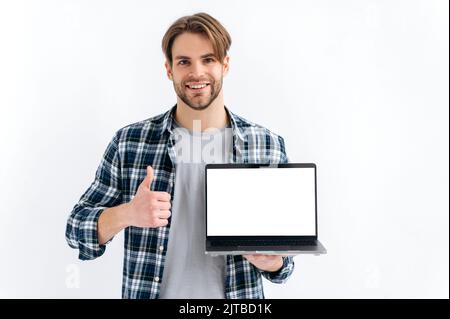 Positive confident caucasian guy in casual wear, freelancer or student, holding an open laptop with empty mock-up space, stand on white isolated background, looks at camera, smiles, shows thumb-up Stock Photo