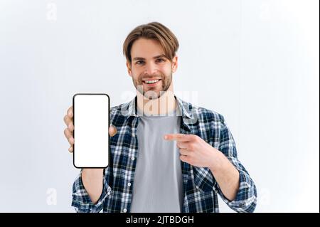 Attractive positive caucasian young man, looks at camera, hold in hand smartphone with white empty mock-up screen for presentation and points finger at it, stand on isolated white background, smiles Stock Photo