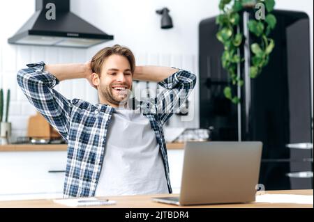 Calm satisfied caucasian guy, freelancer, working from home, sits in the kitchen at workplace, takes a break from work, puts his hands behind his head, looks to the screen, glad with a project, smiles Stock Photo