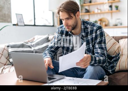 Focused caucasian young man in stylish casual clothes, freelancer, web designer, sitting in living room on the sofa, working on a laptop on a project, analyze documents, looking at the screen intently Stock Photo