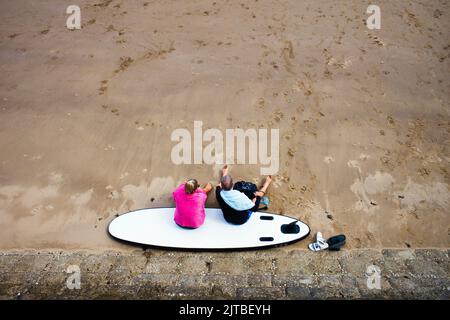 Looking down on an older couple sitting on a surfboard on a sandy beach Stock Photo