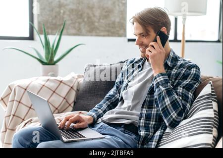 Happy busy caucasian guy in stylish clothes, spending time at home, sits on the sofa in the living room, talking by smartphone with friend, browsing the internet in a laptop, social networks, smile Stock Photo