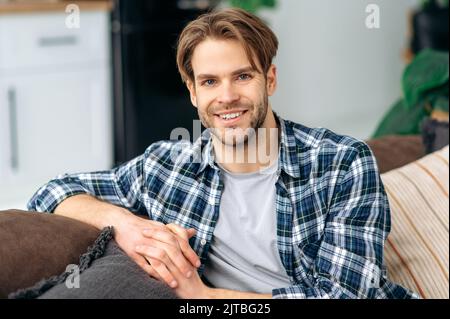 Portrait of confident handsome blue eyed caucasian young man, in casual stylish clothes, sitting at home in cozy living room on the background of the kitchen, looks at the camera, smiling friendly Stock Photo