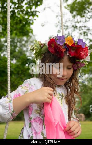 A contemplative girl with long hair wears a flower crown with red poppies, blue campanula, and leaves, while sitting on an outdoor swing. Stock Photo