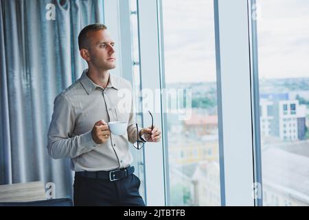 Modern office businessman or manager drinking coffee by the window, smiling. Portrait of successful IT engineer programmer wearing glasses working in Stock Photo