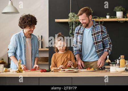 Little boy putting vegetables on bread, helping his parents to cook sandwiches at table in domestic kitchen Stock Photo