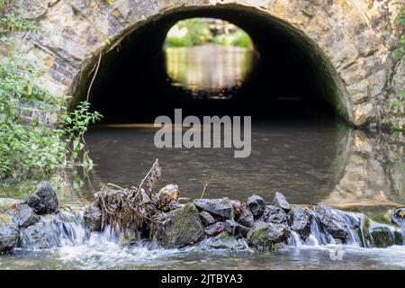 close-up of a chalk stream river flowing out of a stone arched tunnel and over a small stone waterfall Stock Photo