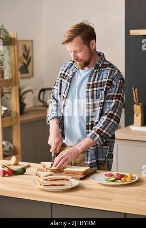 Young man preparing sandwiches with vegetables at wooden table in kitchen Stock Photo