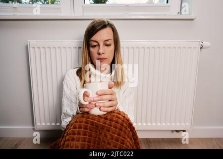 Worried sad woman sits under blanket near heating radiator with cup of tea, Rising costs in private households for gas bill due to inflation and war, Energy crisis Stock Photo