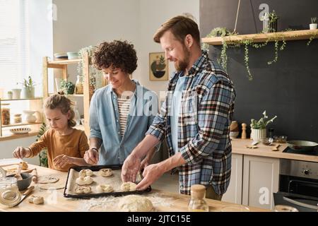 Parents with child preparing homemade buns together, they putting them on tray for baking Stock Photo