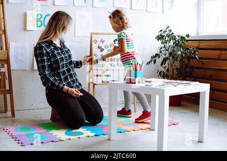 Teacher and girl do exercise on magnetic board. Child learn english alphabet and numbers in kindergarten. Young woman and kindergartener learn to read Stock Photo